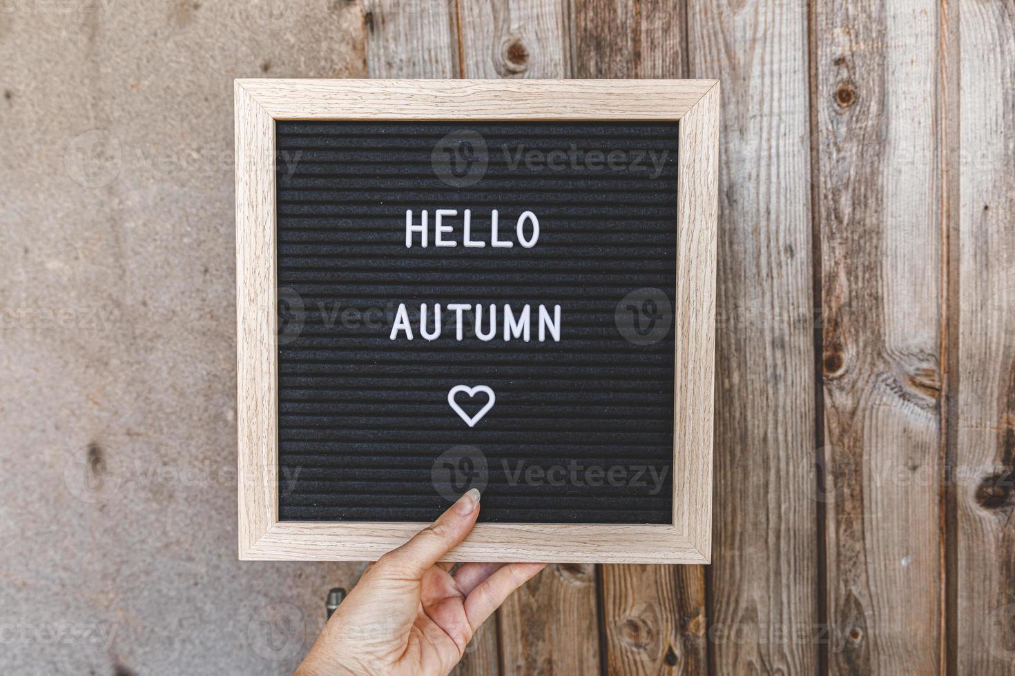 Autumnal Background. Woman hand holding black letter board with text phrase Hello Autumn on wooden palnks backdrop. Thanksgiving banner. Hygge mood, cold weather concept photo