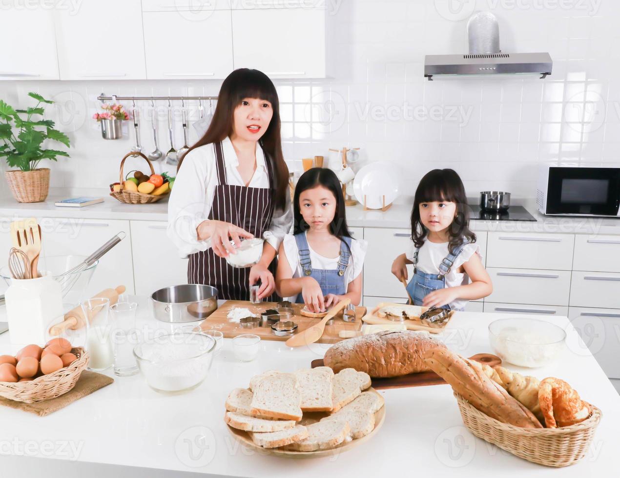 mamá asiática y dos hijas pequeñas con delantal cocinando juntos en la cocina foto