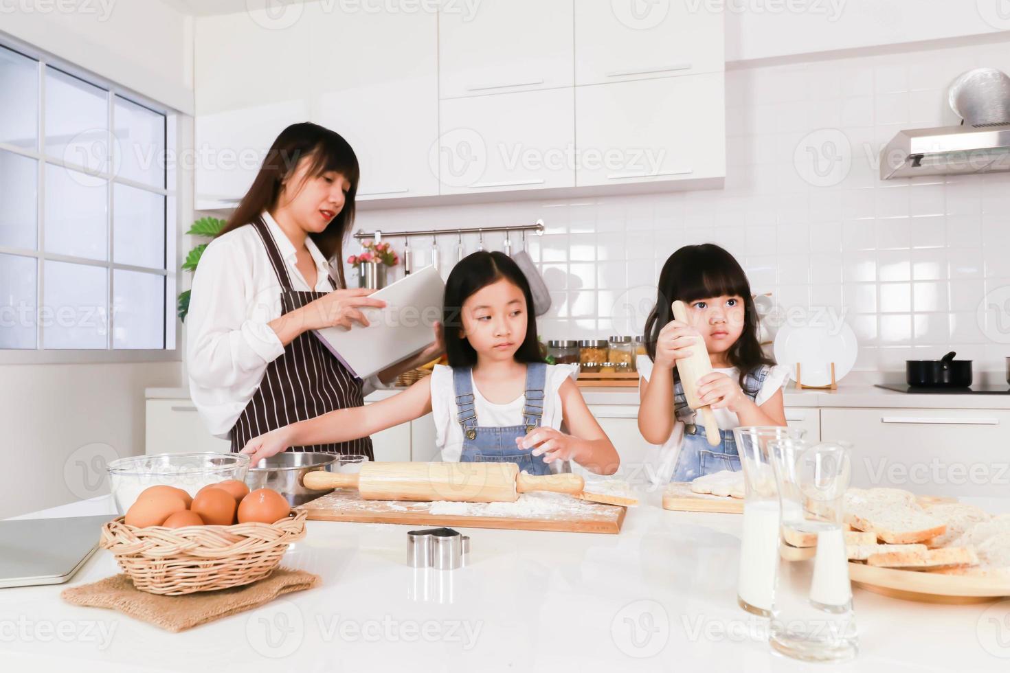 familia asiática cocinando juntos mamá leyendo un libro y dos hijas preparando un horneado foto