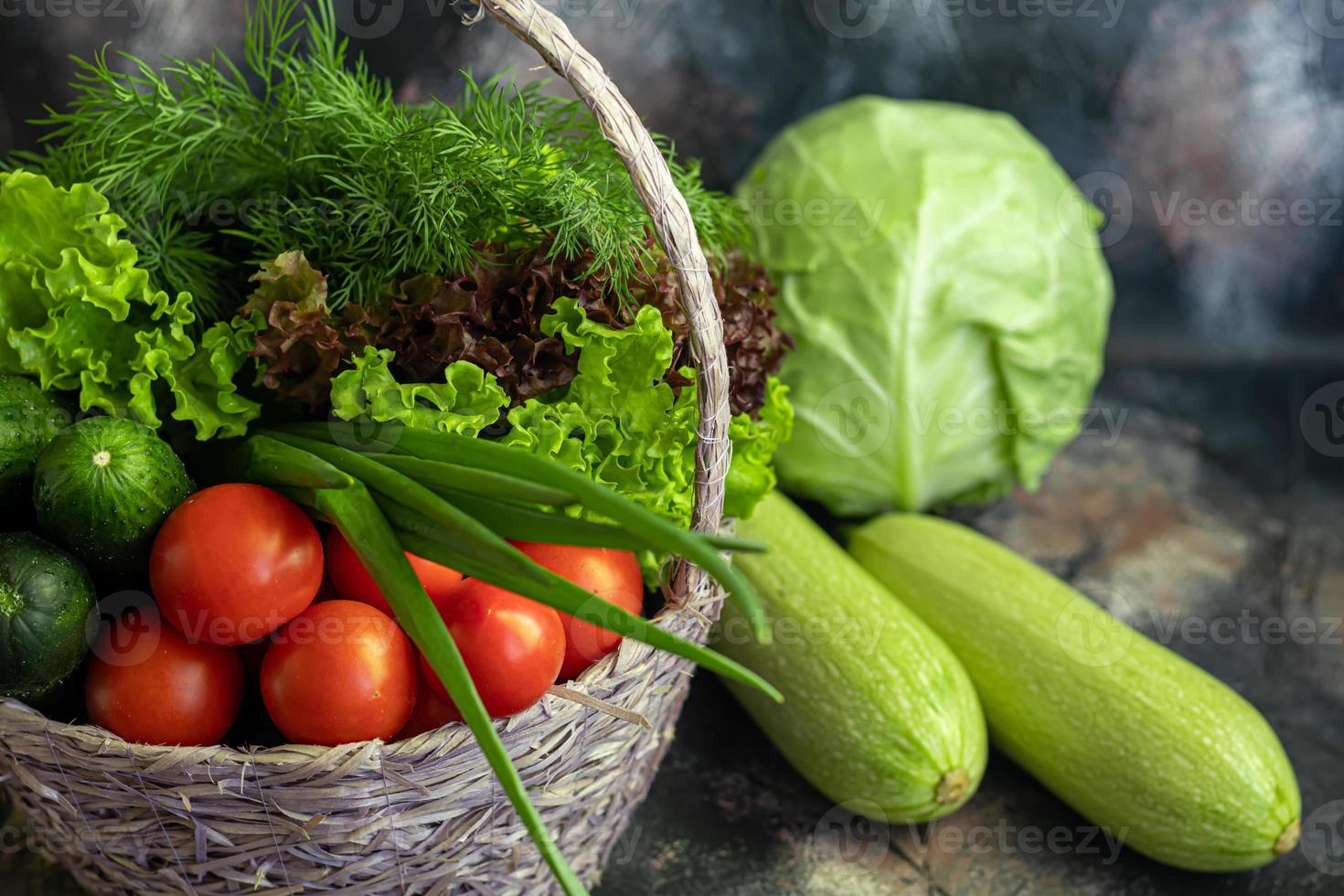 Fresh vegetables for salad in a basket. Tomatoes and cucumbers with zucchini and cabbage with dill. Spring harvest, benefits and vitamins. On a dark background. photo