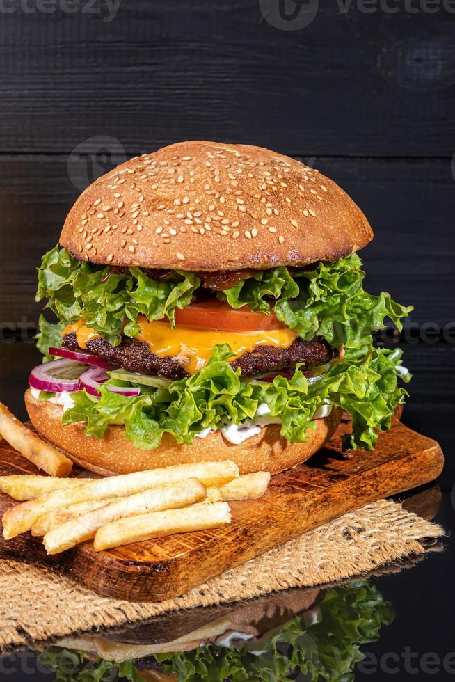 Big burger with beef patty, cheese, cucumber and lettuce. French fries on a wooden board. On a dark background. photo