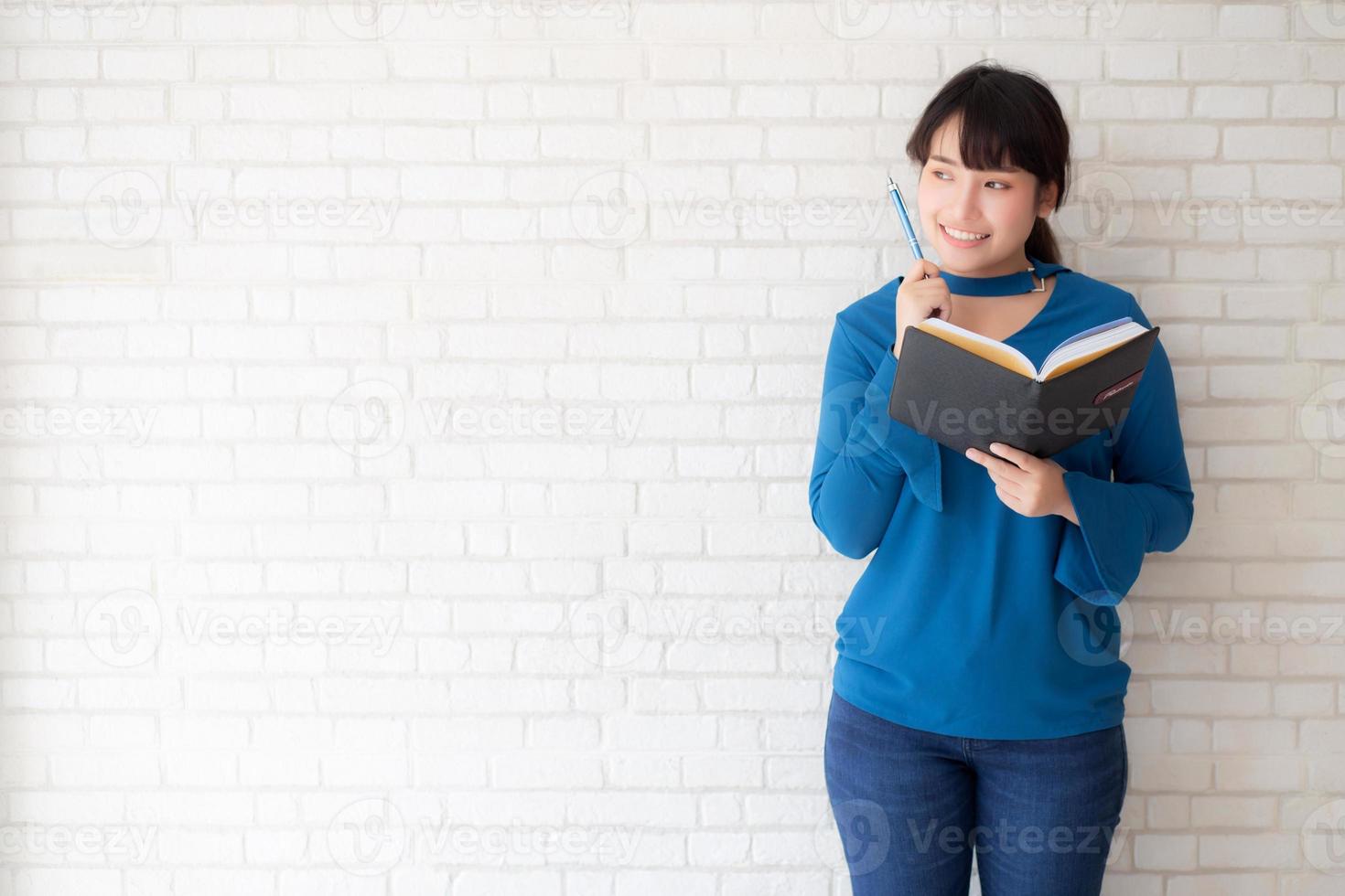 Beautiful asian woman smiling standing thinking and writing notebook on concrete cement white background at home, girl homework on book, education and lifestyle concept. photo