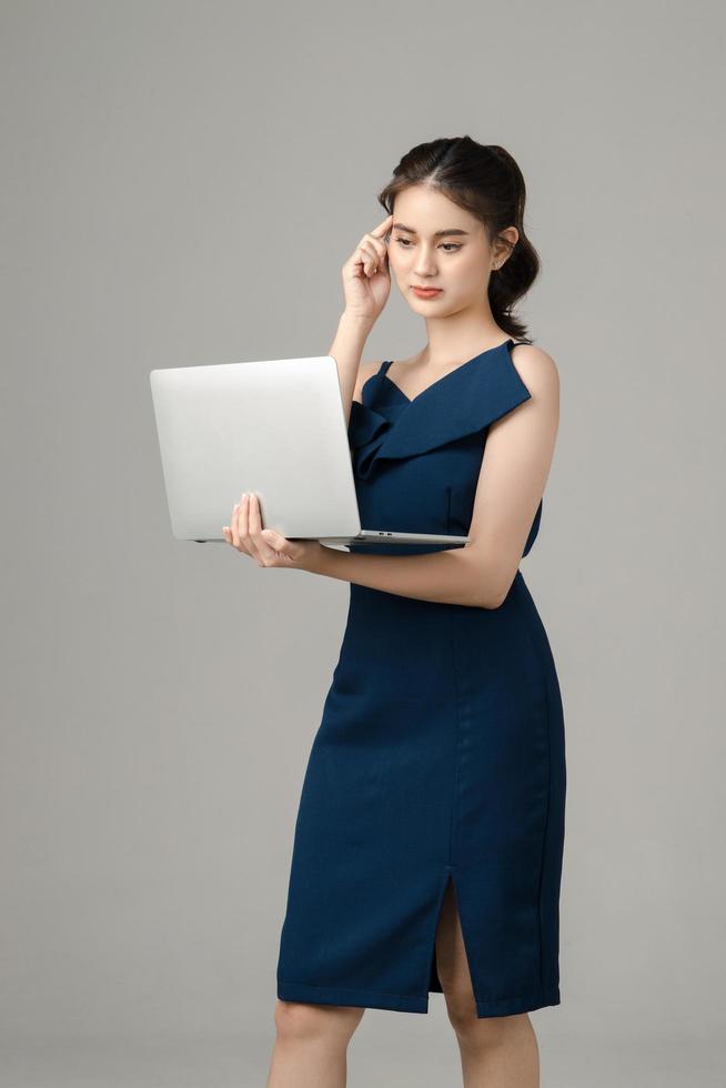 Young serious Asian business woman holding laptop and thinking on gray background. Portrait of pretty girl in studio. Small business SME, freelance online, e-commerce Concept. photo