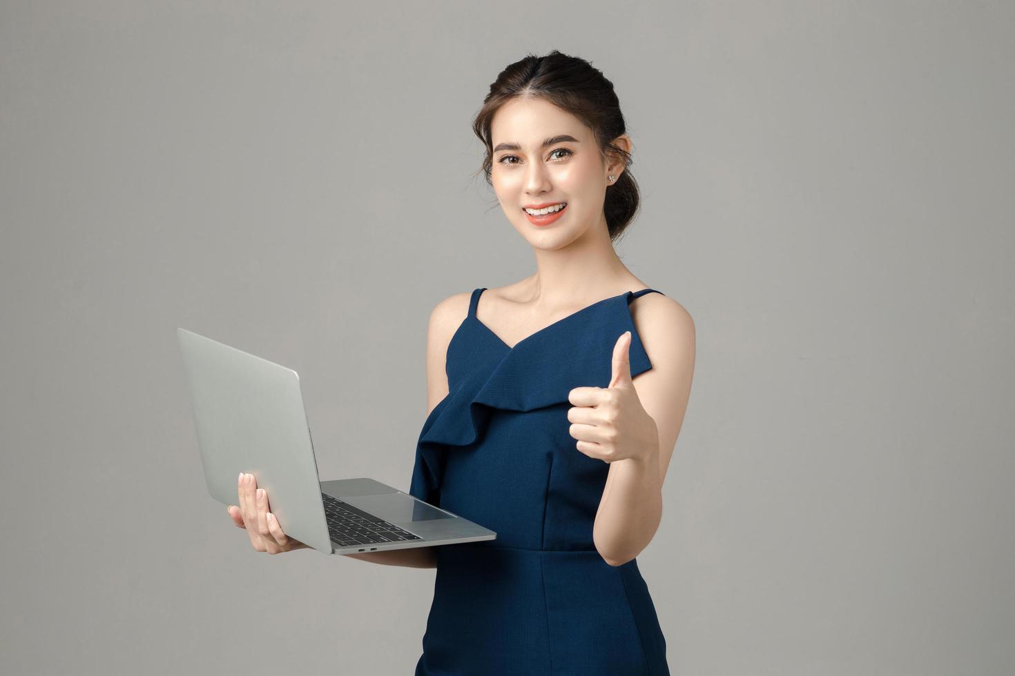 joven y enérgica mujer de negocios asiática sosteniendo una laptop mostrando el pulgar hacia arriba sobre fondo gris. retrato de niña bonita en estudio. pyme de pequeñas empresas, autónomo en línea, concepto de comercio electrónico. foto