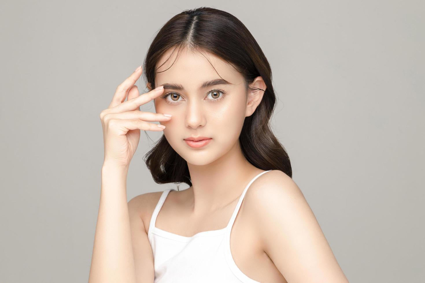 hermosa joven modelo asiática toca su cara con una piel limpia y fresca perfecta sobre fondo gris. cuidado facial, tratamiento facial, cosmetología, cirugía plástica, hermoso retrato de niña en estudio. foto