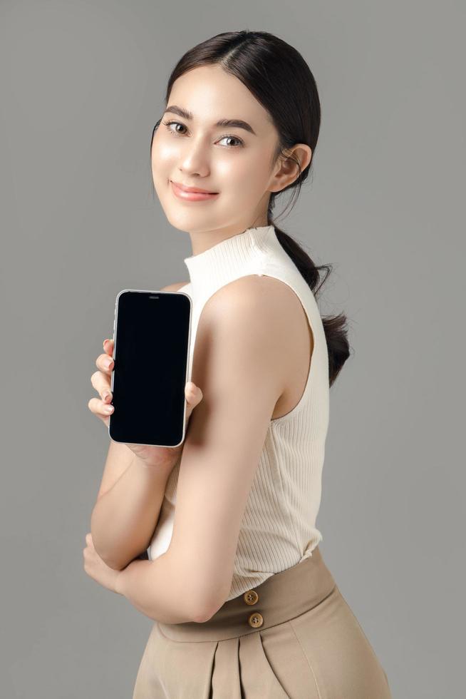 mujer asiática segura de sí misma sosteniendo un teléfono que muestra una pantalla en blanco y mirando una cámara aislada en un fondo gris. retrato de una chica hermosa en el estudio. foto