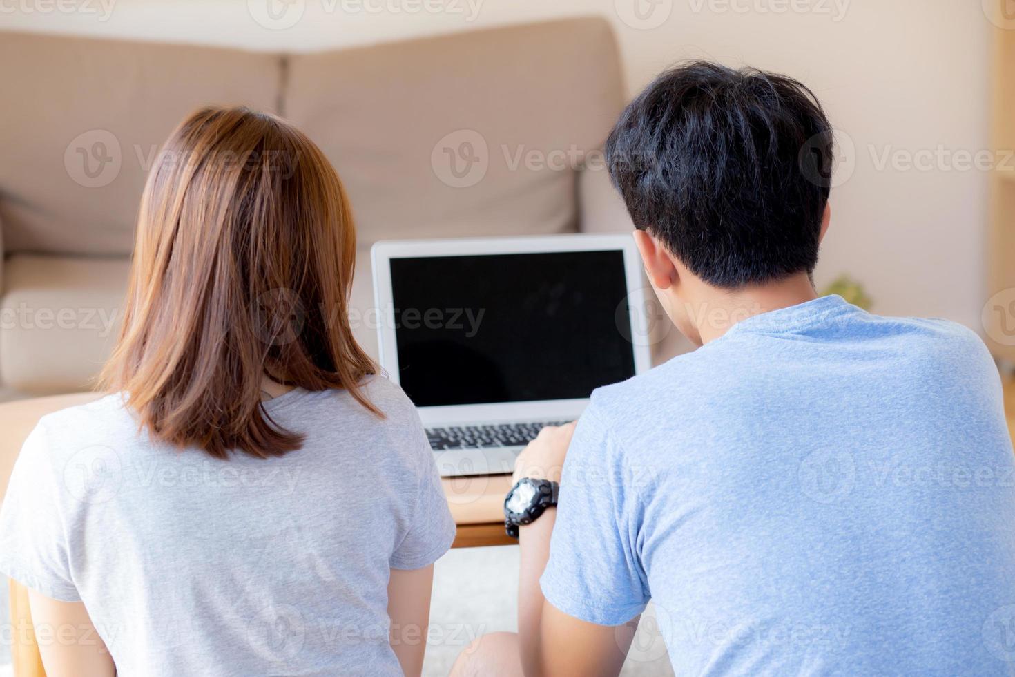 Back view of young asian couple working laptop with blank screen display, family planning and searching content together, man and woman looking computer, business and communication concept. photo