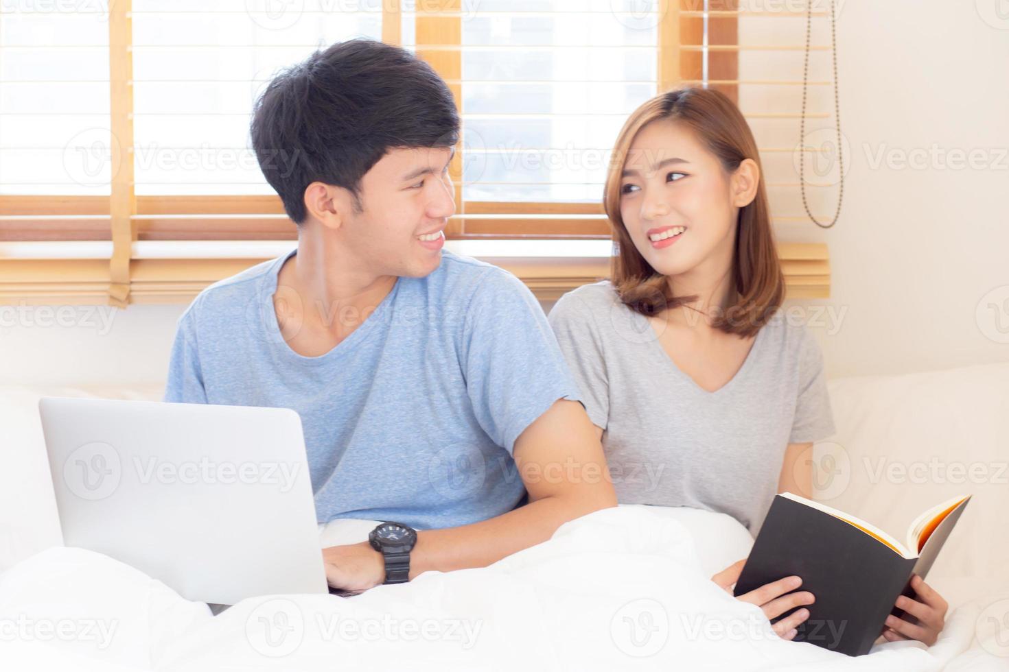 Asian young couple with man using laptop computer and searching internet and woman reading notebook on bed at bedroom with happy and relax, communication and business concept. photo