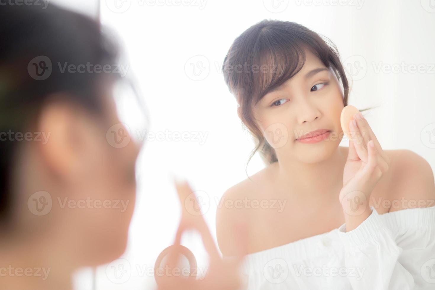 Portrait beautiful young asian woman applying powder puff at cheek makeup of cosmetic looking mirror, beauty of girl with skin face smile in the bedroom, health and wellness concept. photo