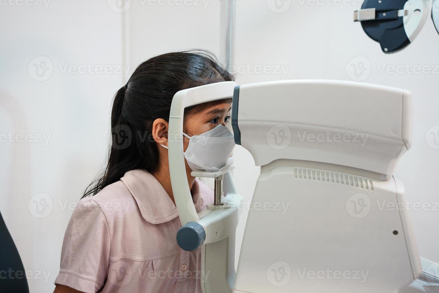 Ophthalmologist examining the eyes of an Asian girl in a clinic. They wear protective face masks. photo