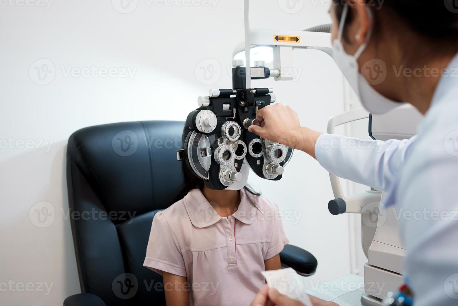 Ophthalmologist examining the eyes of an Asian girl in a clinic. They wear protective face masks. photo