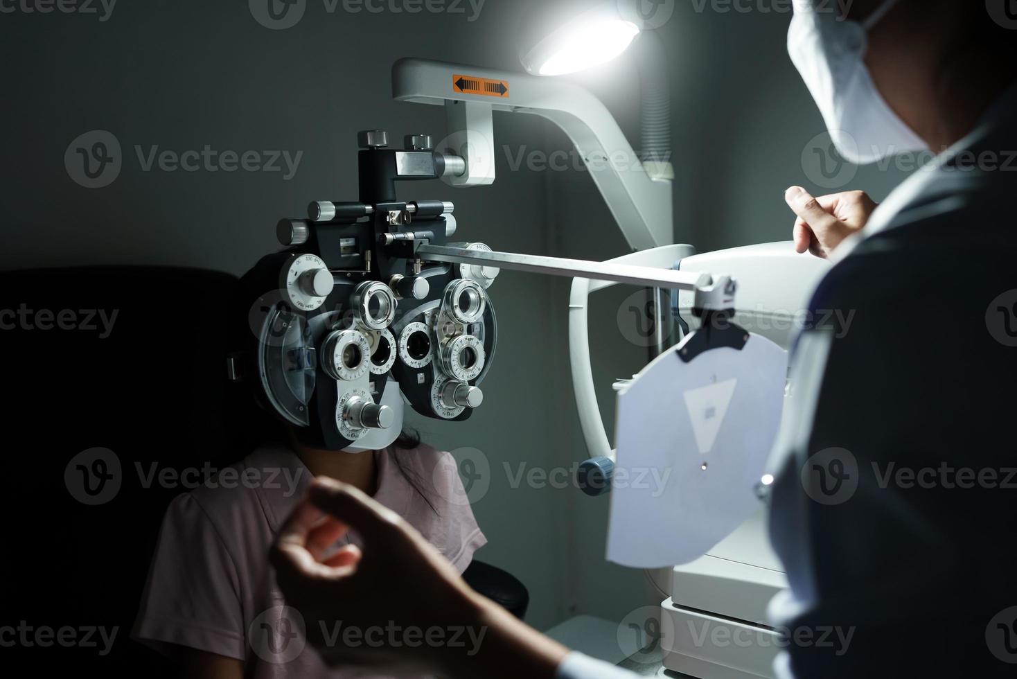 Ophthalmologist examining the eyes of an Asian girl in a clinic. They wear protective face masks. photo