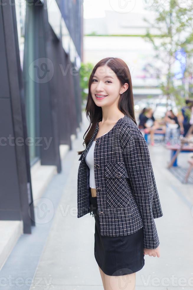 Portrait of a beautiful, long-haired Asian female in a black pattern coat with braces on teeth walking and smiling outdoors in the city. photo