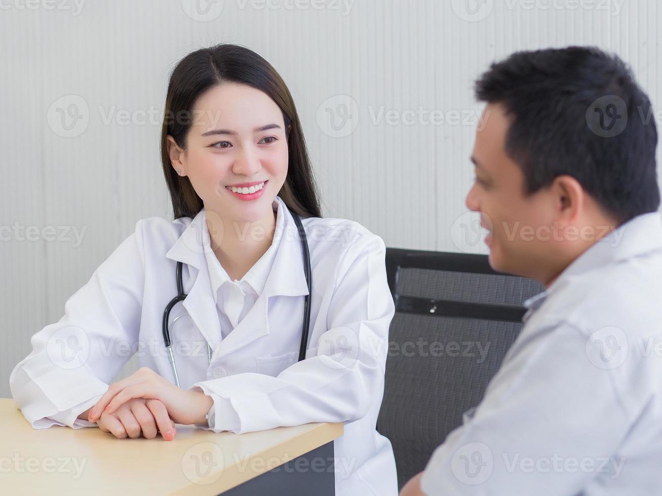 Professional beautiful young Asian female doctor talking with a man patient about his pain and symptom while they put on a face mask to Thermometer on table at the examination room in hospital. photo