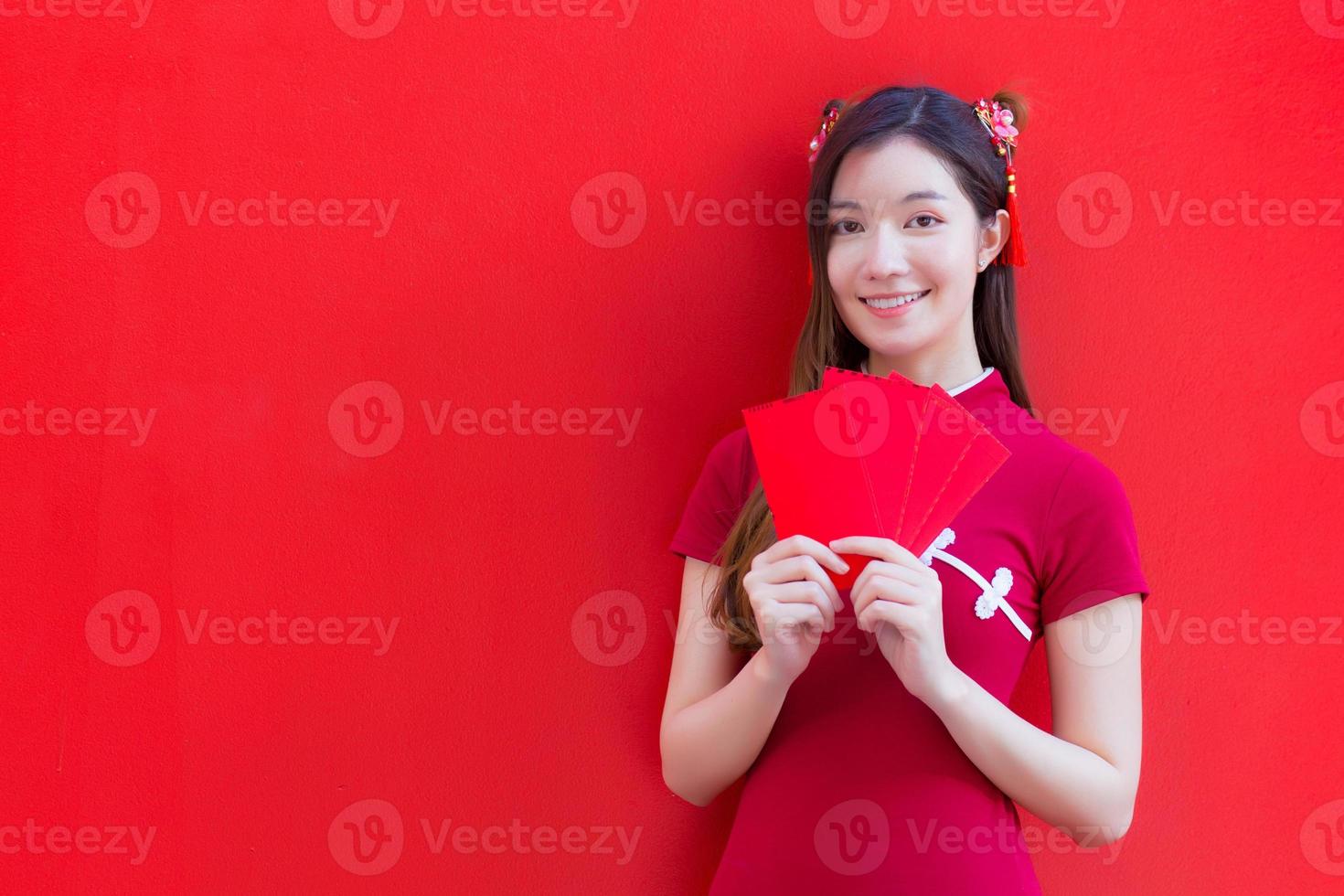 Beautiful Asian female wears a red cheongsam and holds red envelopes while looks at to camera and smiles happily with the red background. photo