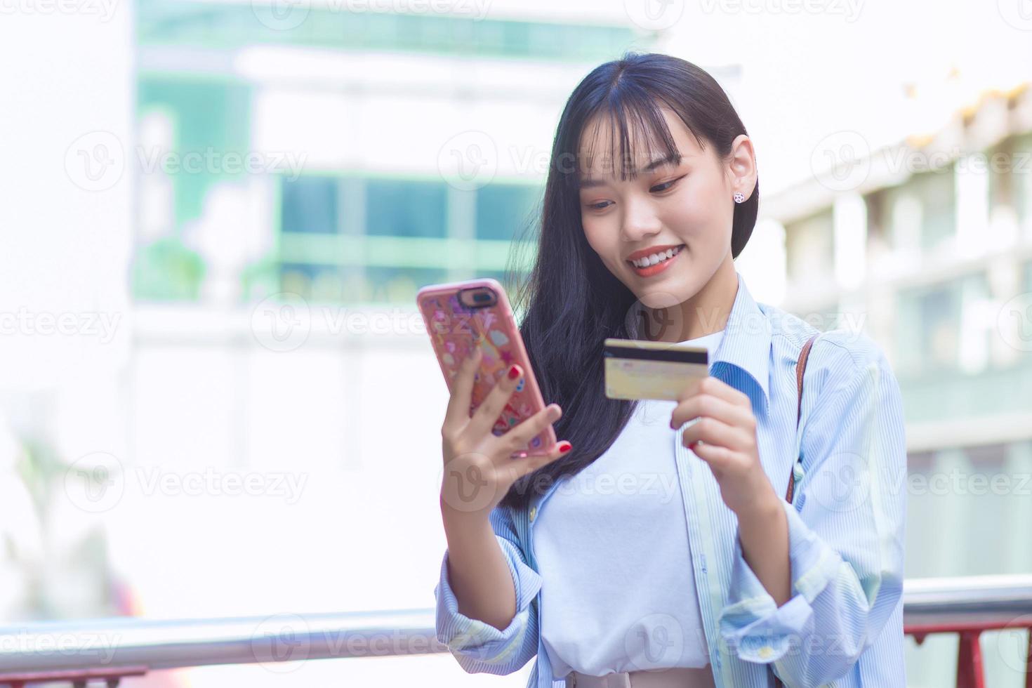 Professional Asian business woman in a blue shirt smiles happily successful outdoors in the city while holds smartphone and credit card among the building as a background. photo