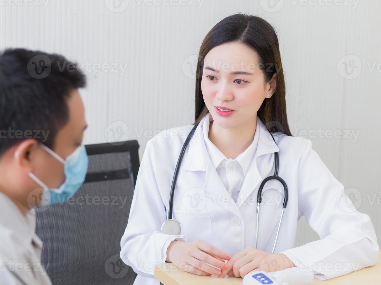 Professional beautiful young Asian female doctor talking with a man patient about his pain and symptom while they put on a face mask to prevent Coronavirus disease and Thermometer on table.3 photo