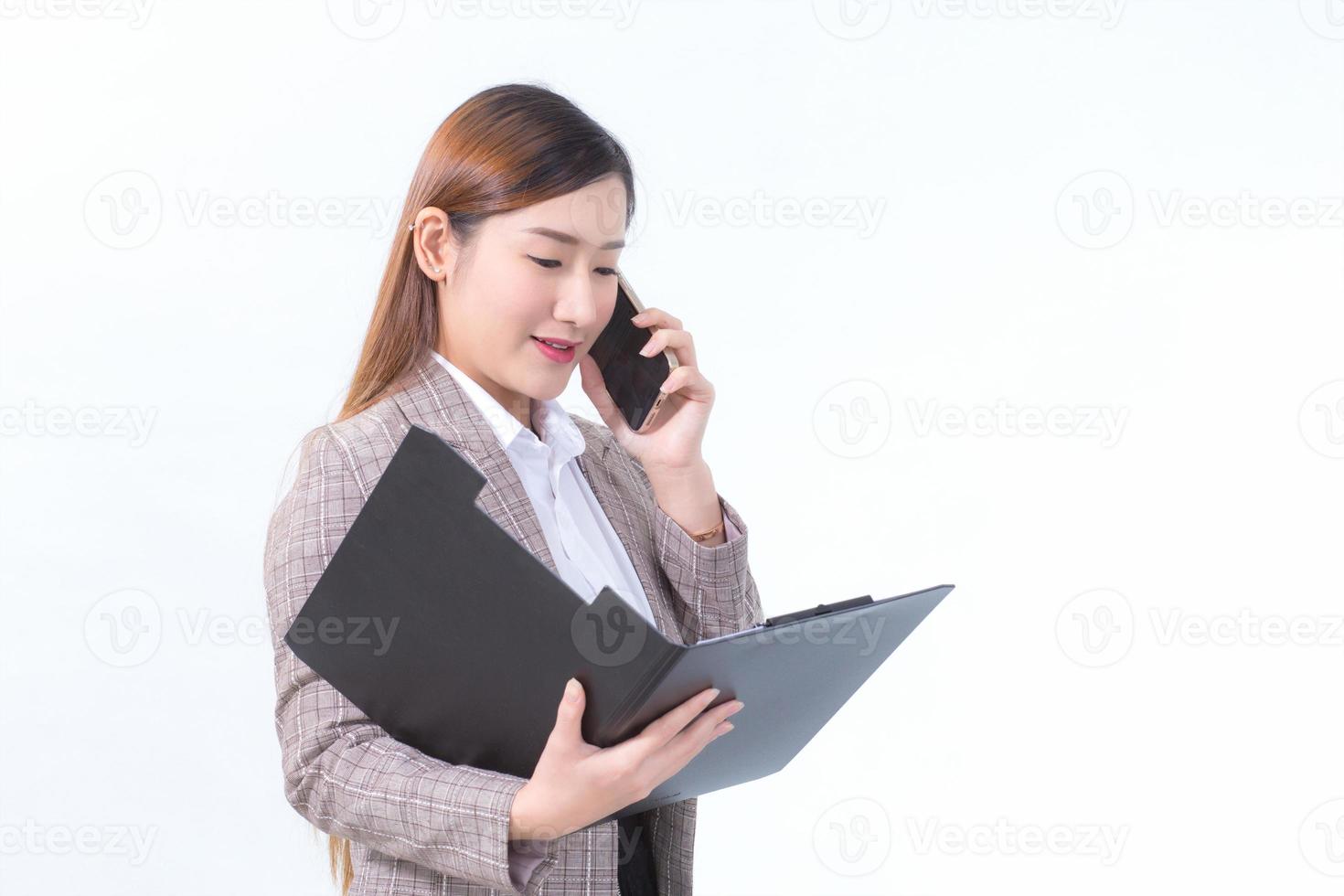 una trabajadora asiática con traje formal y camisa blanca llama por teléfono y abre un archivo de documento o un portapapeles para verificar los datos. foto