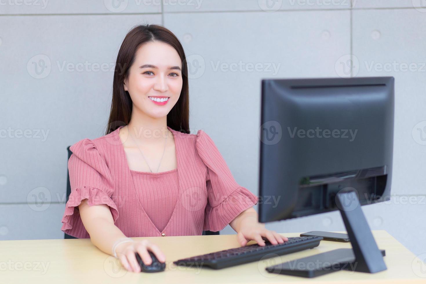 Professional young Asian business woman who wears pink dress is working while she sits  and smiles in an office typing on a keyboard and mouse. photo