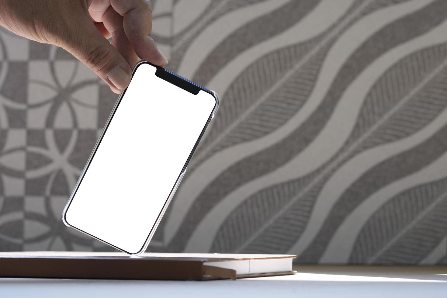 Close up man's hand showing smartphone with blank screen in leaning gesture on book with blurred abstract pattern on brown tile wall background photo