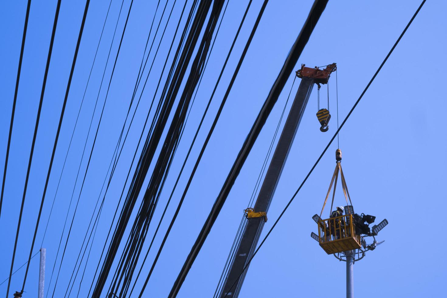 vista de ángulo bajo del electricista en la grúa que repara el sistema eléctrico de la torre del foco con muchas líneas eléctricas contra el fondo del cielo azul foto