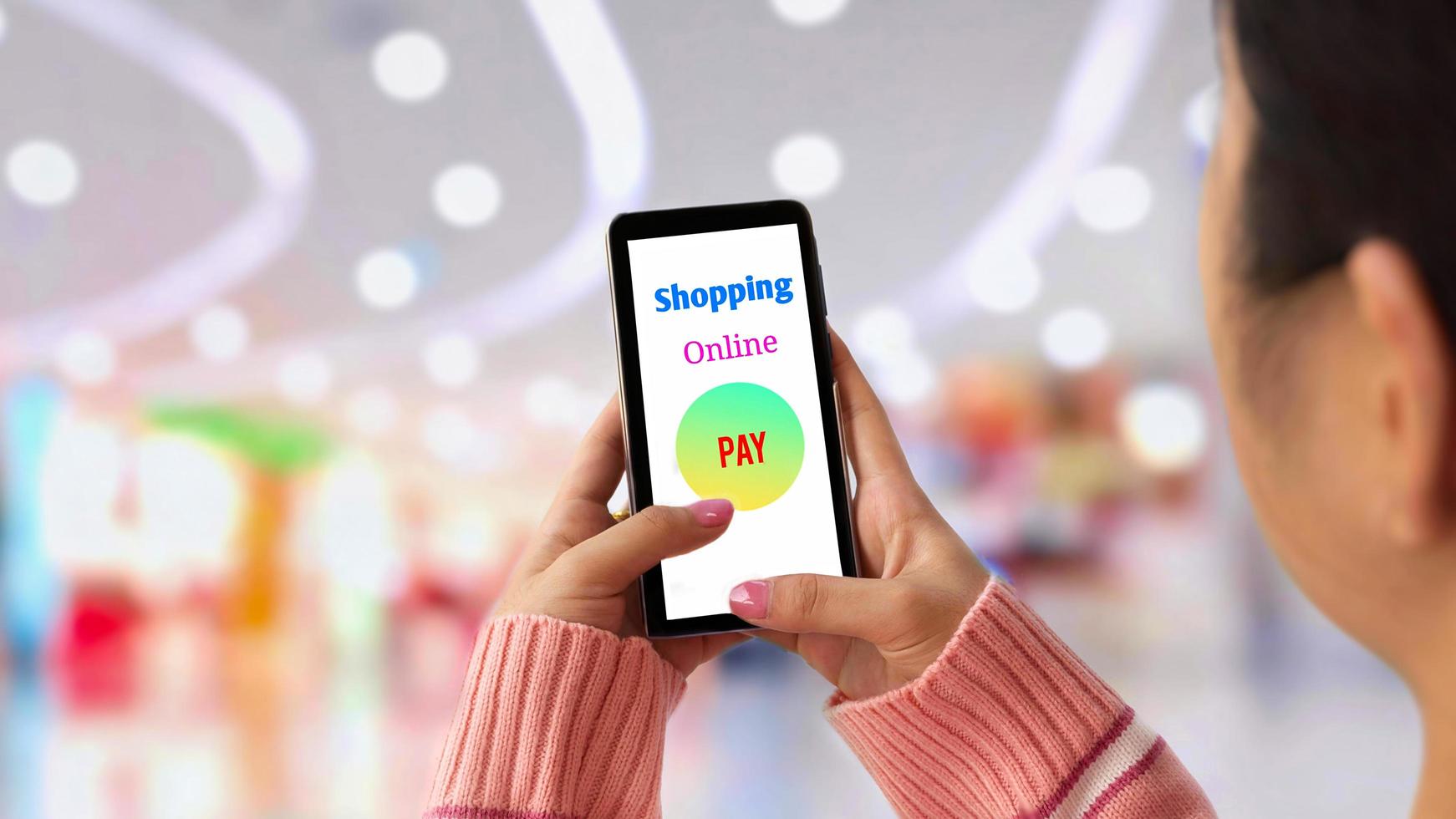 Selective focus at young female hand in pink knitted sweater using application on smartphone to shopping online payment with blurred shopping mall background photo