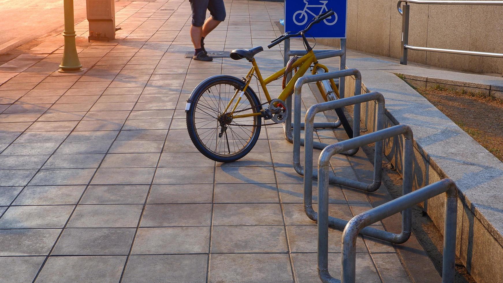 Selective focus at the old bicycle parked in row of stand bike parking rack with blue bicycle parking sign and low section of man walking on roadside pavement floor photo