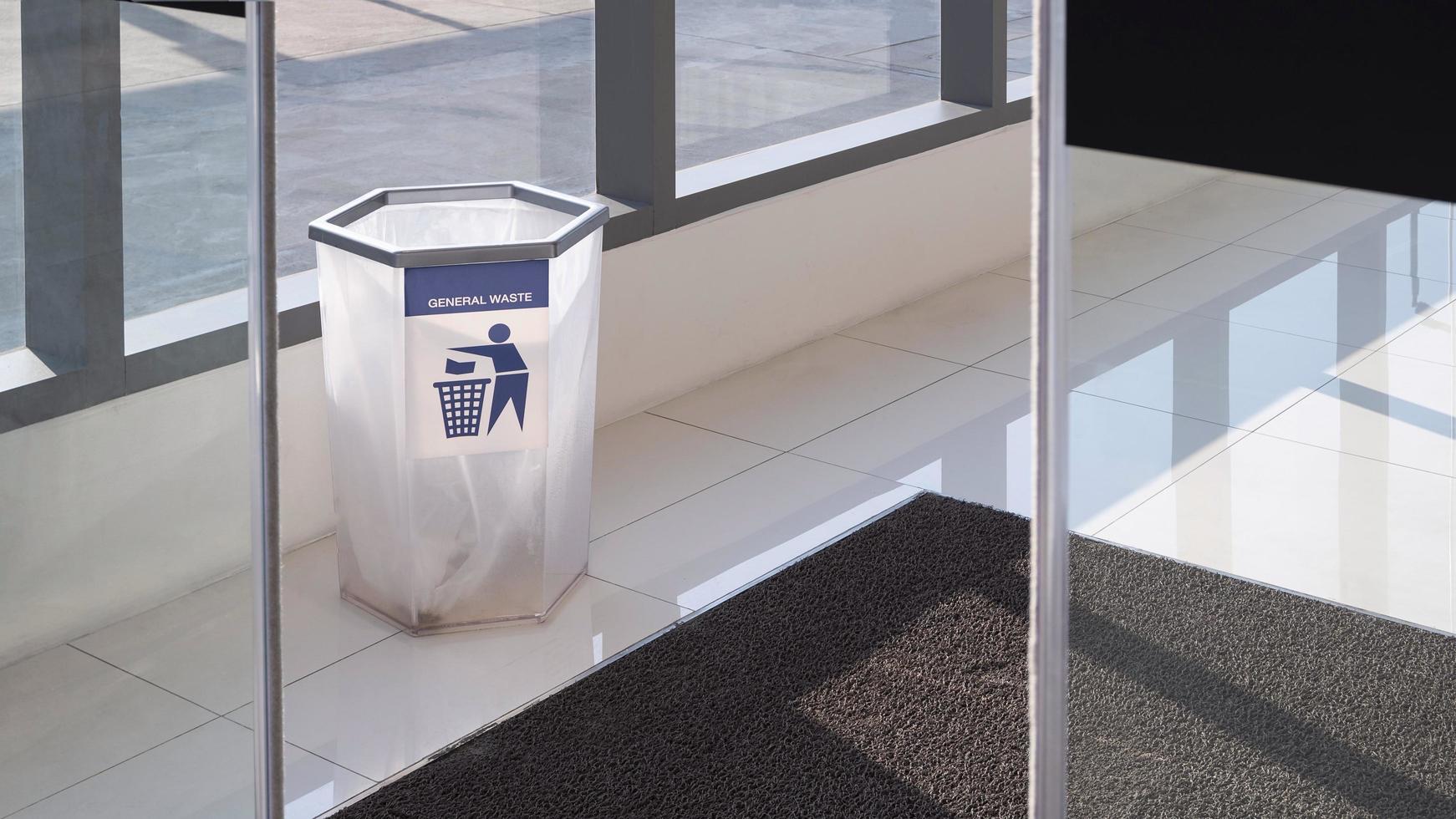 Focus at transparent hexagonal shape trash bin with black rubber doormat on white tile floor of entrance way of shopping mall with blurred automatic glass sliding door in foreground photo