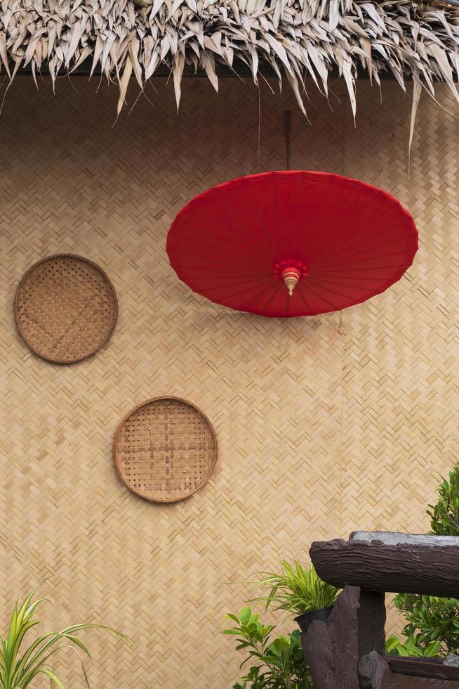 Red paper umbrella with 2 threshing basket on bamboo weave wall of traditional hut with blurred green houseplant decoration on foreground in vintage style and vertical frame photo