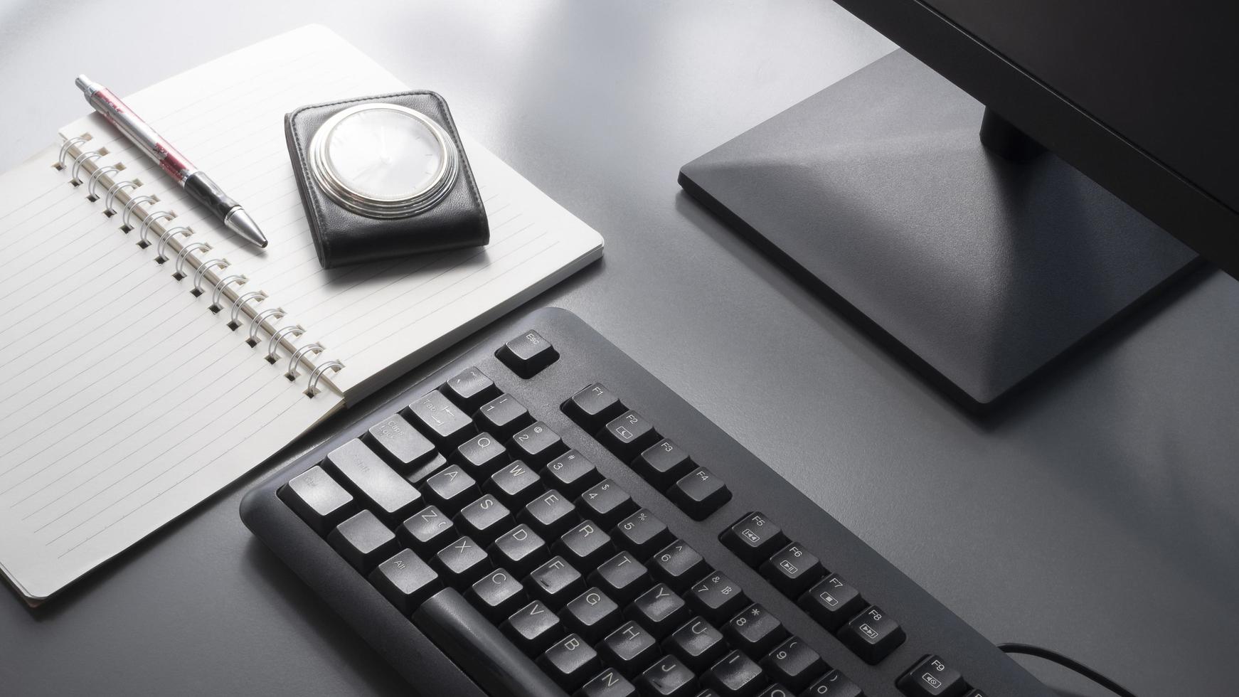 Selective focus at black keyboard with part of computer monitor, ballpoint pen, vintage travel leather case pocket watch and spiral notebook on gray tabletop with sunlight and shadow on surface photo