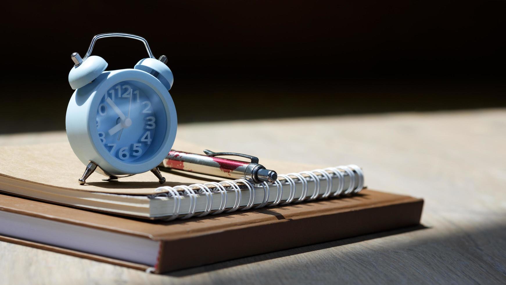 Selective focus at little blue alarm clock with ballpoint pen and kraft paper books with sunlight and shadow on wooden table surface, hobby and relaxing time concept photo