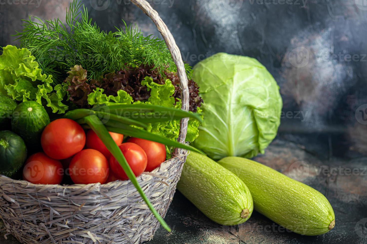 verduras frescas para ensalada en una cesta. tomates y pepinos con calabacín y repollo con eneldo. cosecha de primavera, beneficios y vitaminas. sobre un fondo oscuro. foto