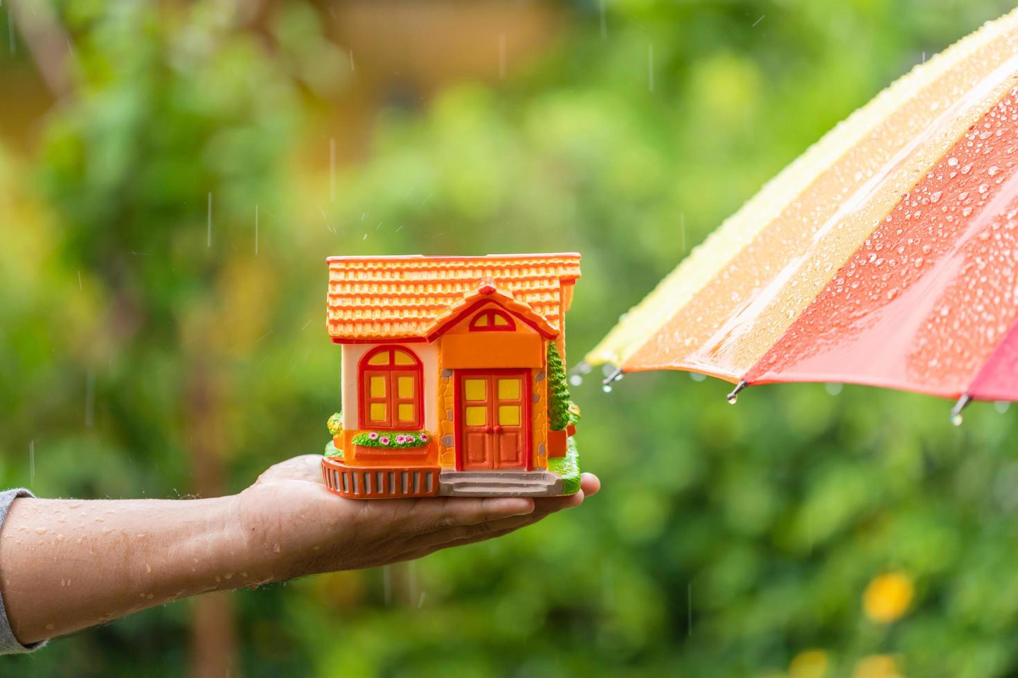 Hand holding piggy bank Home Model beside wet umbrella in rainy days on green nature blur background. Home protect and maintenance in rainy season concept photo