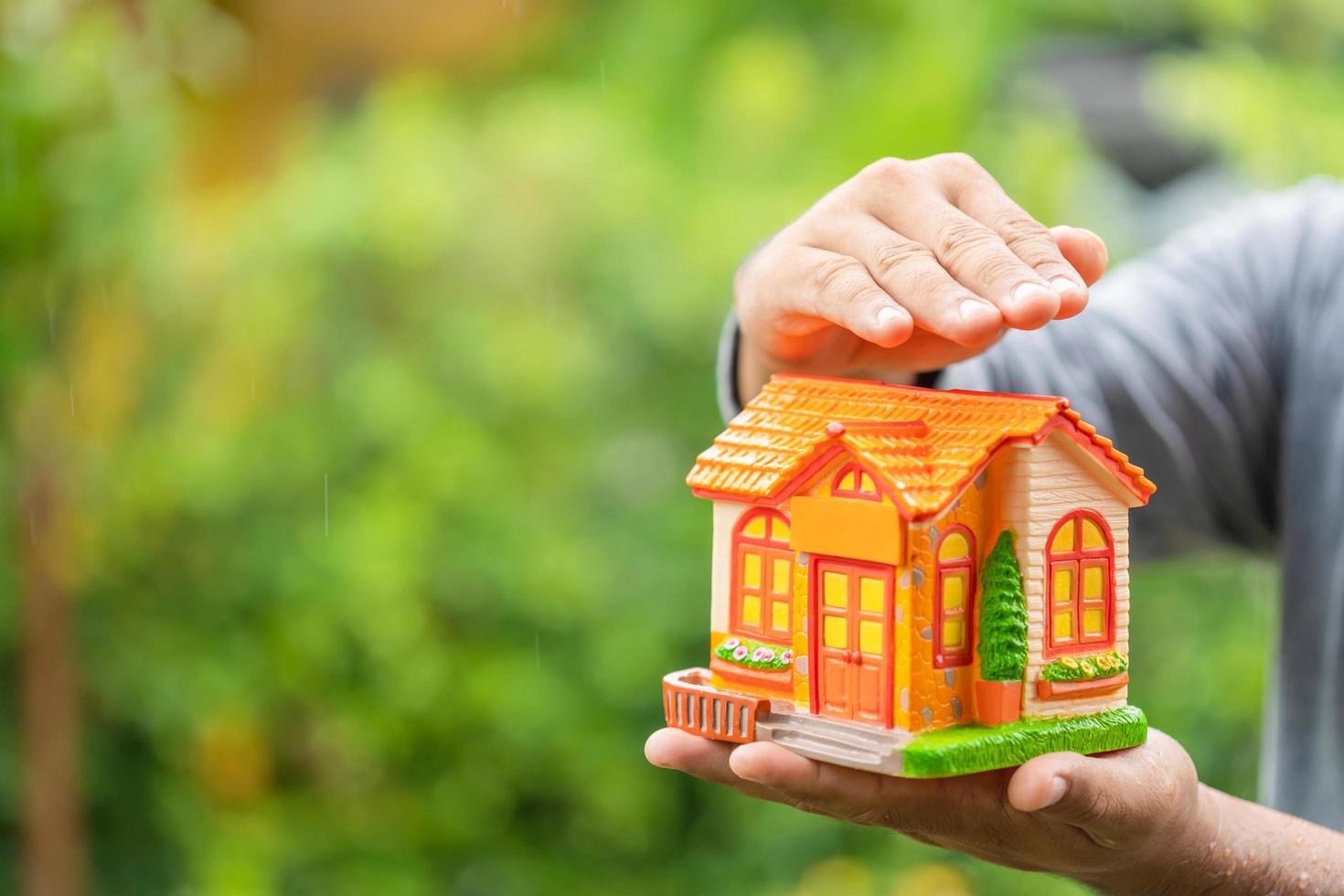 Hand holding piggy bank Home Model in rainy days on green nature blur background. Home protect and maintenance in rainy season concept photo