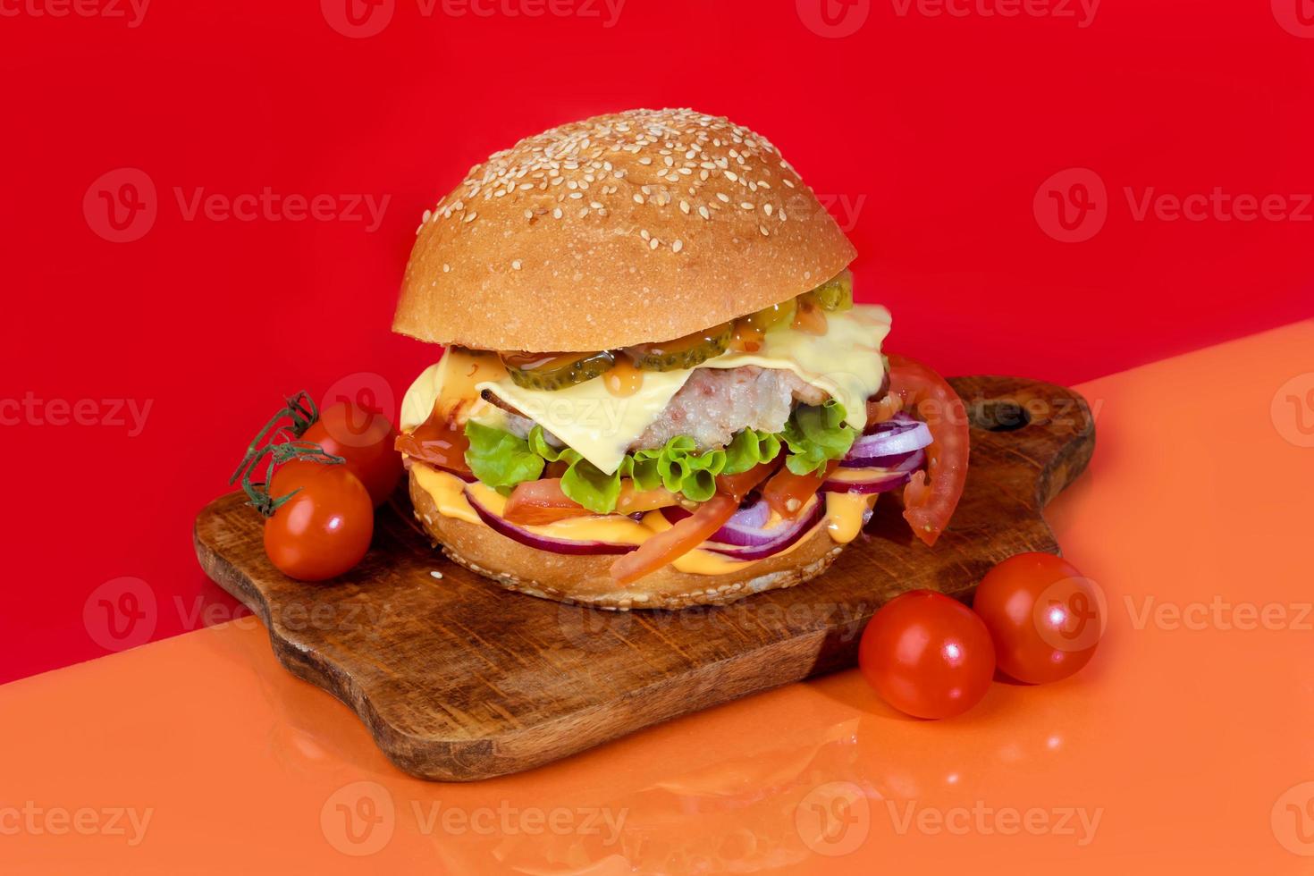 Delicious burger on the board with cutlet, tomatoes and cheese, cucumber and lettuce. On a red background. Fast food. photo