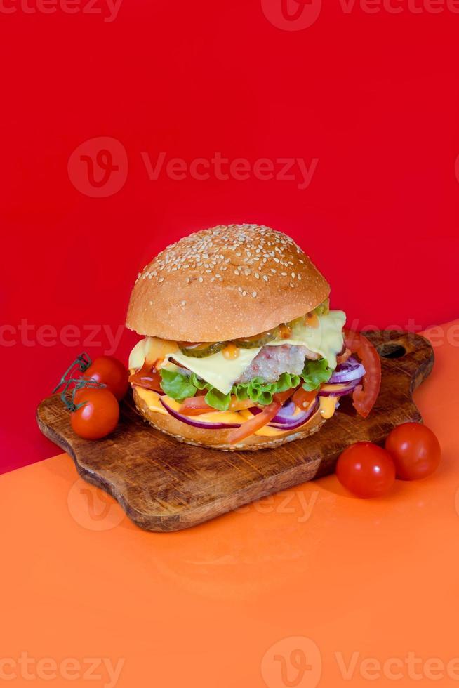 Delicious burger on the board with cutlet, tomatoes and cheese, cucumber and lettuce. On a red background. Fast food. photo