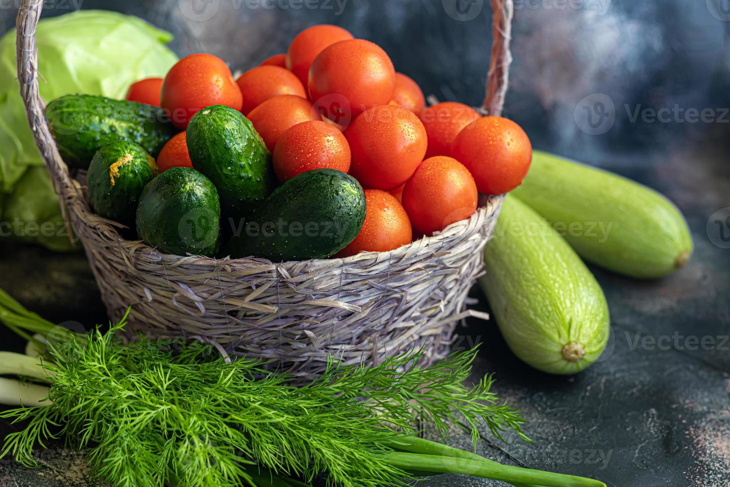 Fresh vegetables for salad in a basket. Tomatoes and cucumbers with zucchini and cabbage with dill. Spring harvest, benefits and vitamins. On a dark background. photo