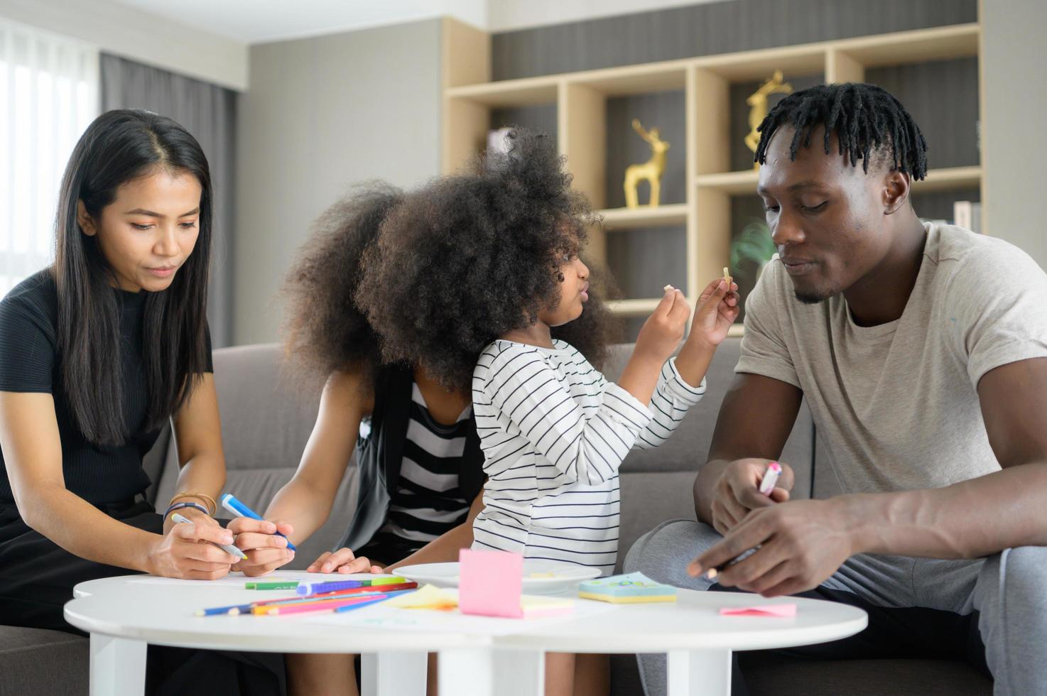 familia asiático-afroamericana relajándose, charlando, pintando y divirtiéndose de vacaciones en la sala de estar de la casa foto