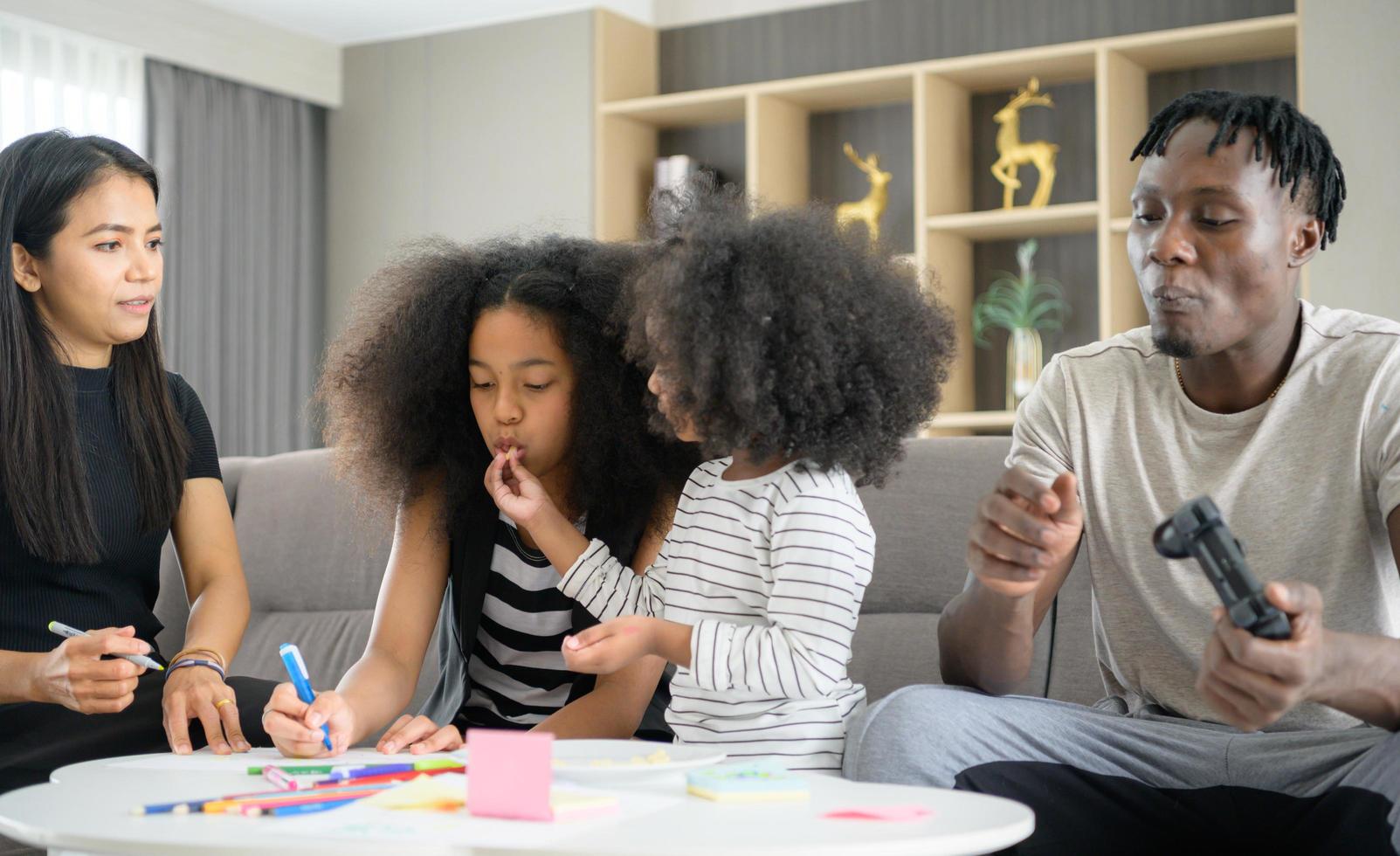 familia asiático-afroamericana relajándose, charlando, pintando y divirtiéndose de vacaciones en la sala de estar de la casa foto