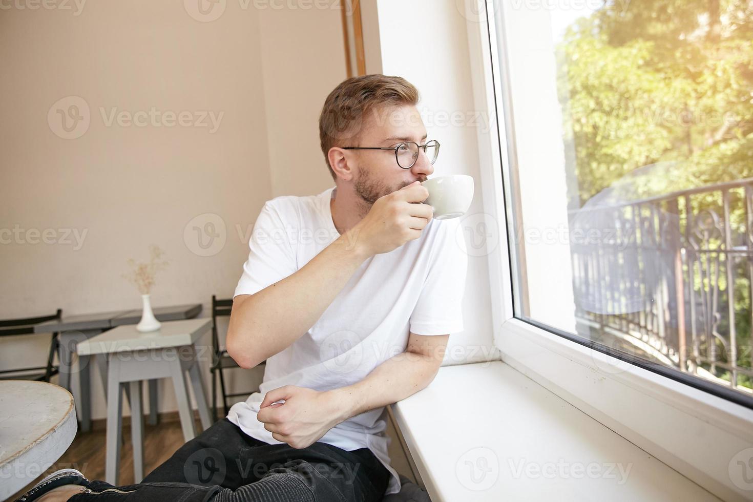 Young short haired attractive man in glasses sitting next to window, drinking coffee and enjoying the view, looking dreamy and meditatively photo