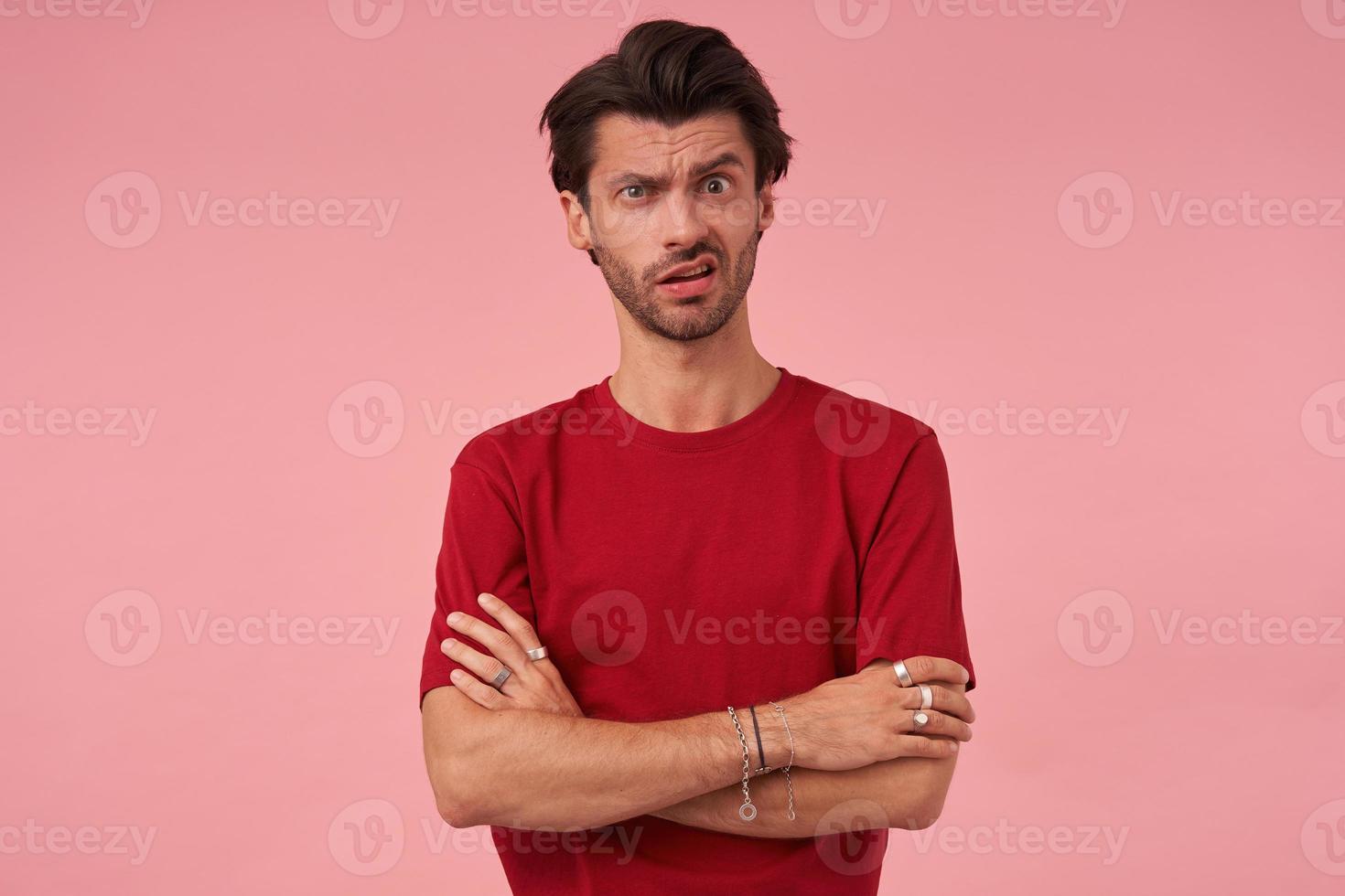 escéptico joven sospechoso con rastrojo y una ceja levantada cuestionando en camiseta roja pensando y de pie con los brazos cruzados sobre fondo rosa foto