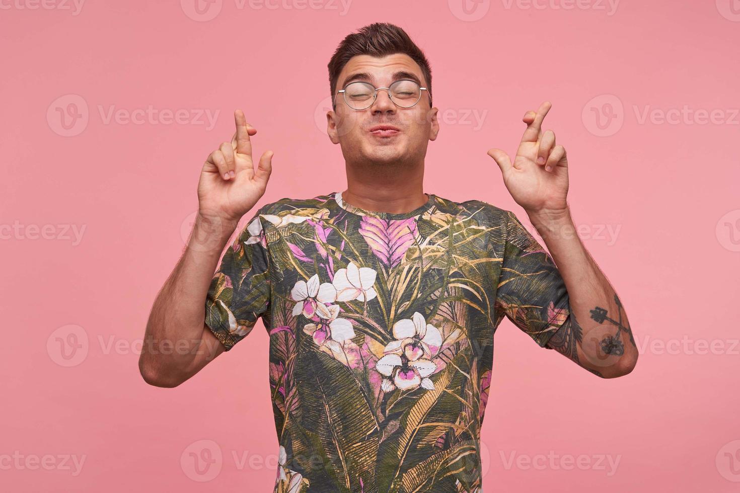 Close-up of positive pretty young man crossing fingers, closing eyes with pleasure and pursing lips, foresee hearing good news, standing over pink background photo