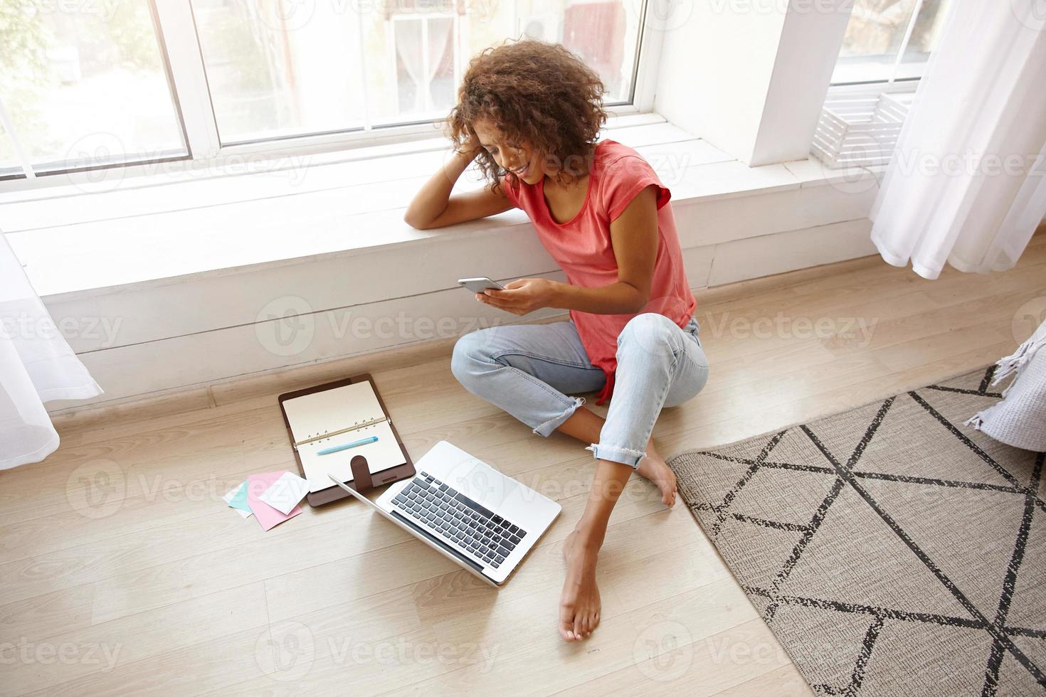 Young good looking female with brown curly hair sitting near wide window and resting her head on hand, working out of office, making break and texting to friend with her smart phone photo