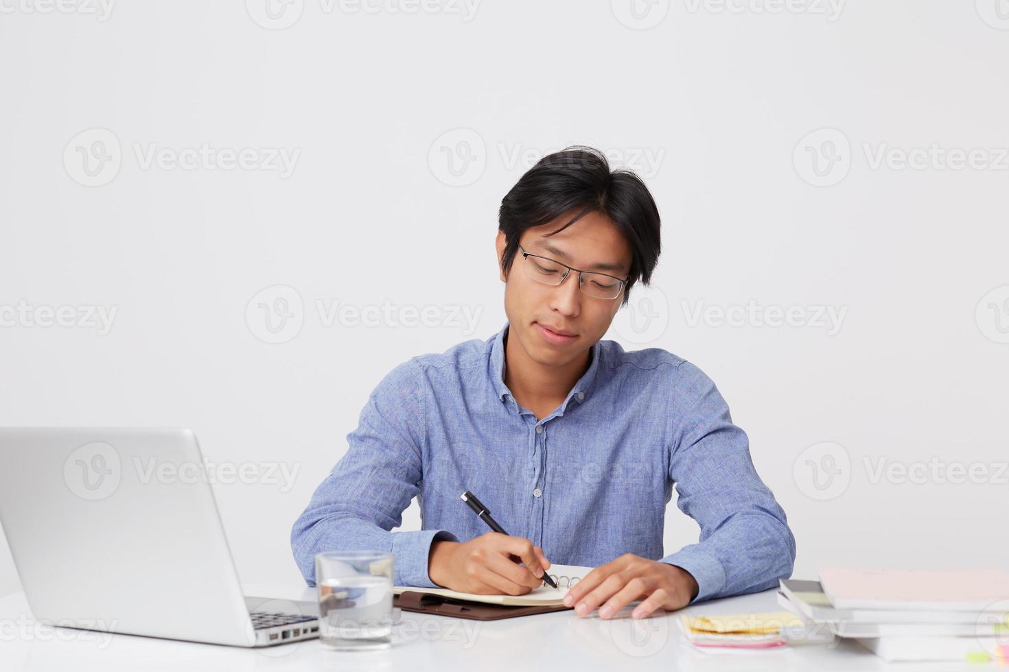 Serious successful asian young business man in glasses working with laptop and writing in notebook at the table over white background photo