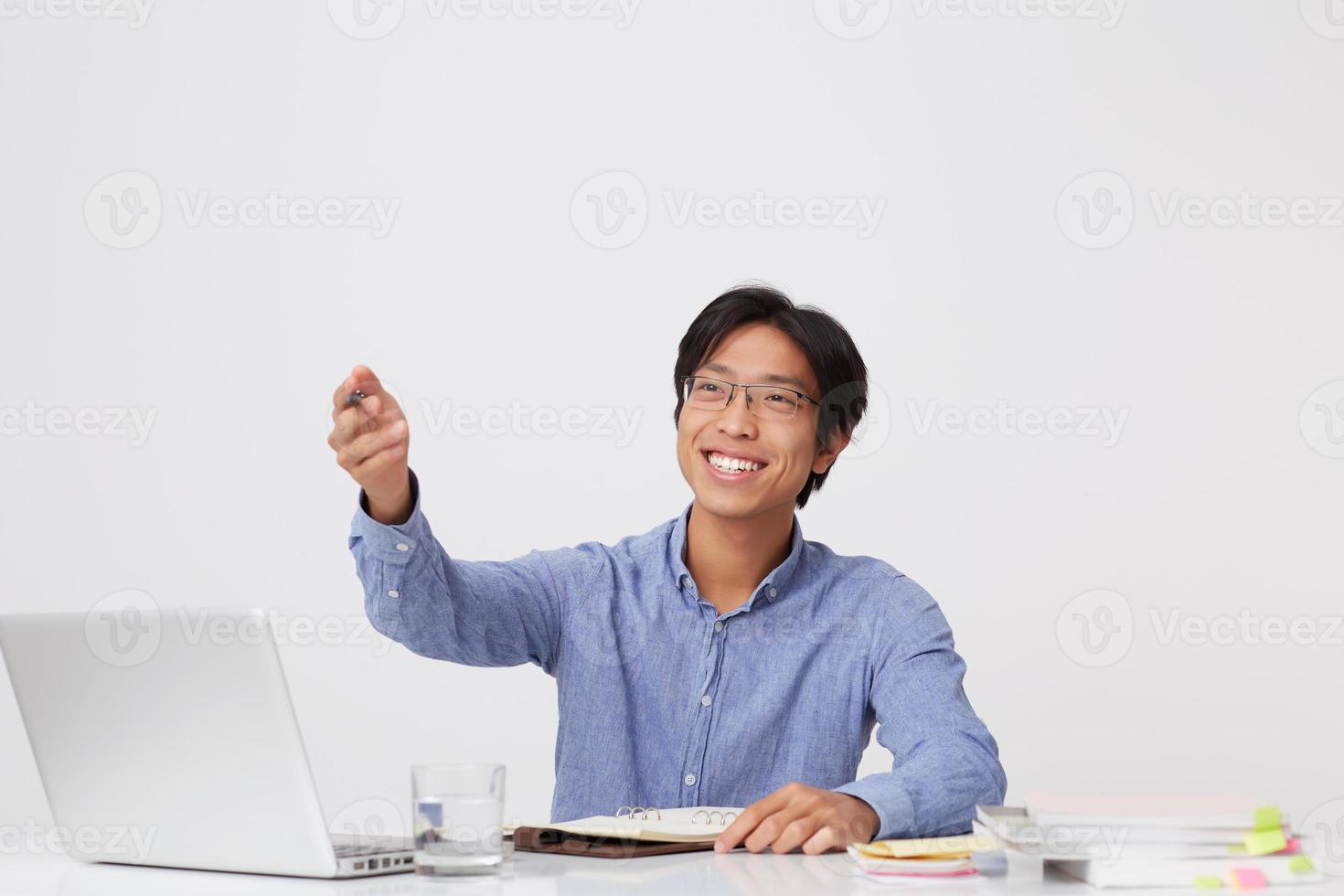 Cheerful attractive asian young business man in glasses writing in notebook and showing to the side by hand at the table over white background photo