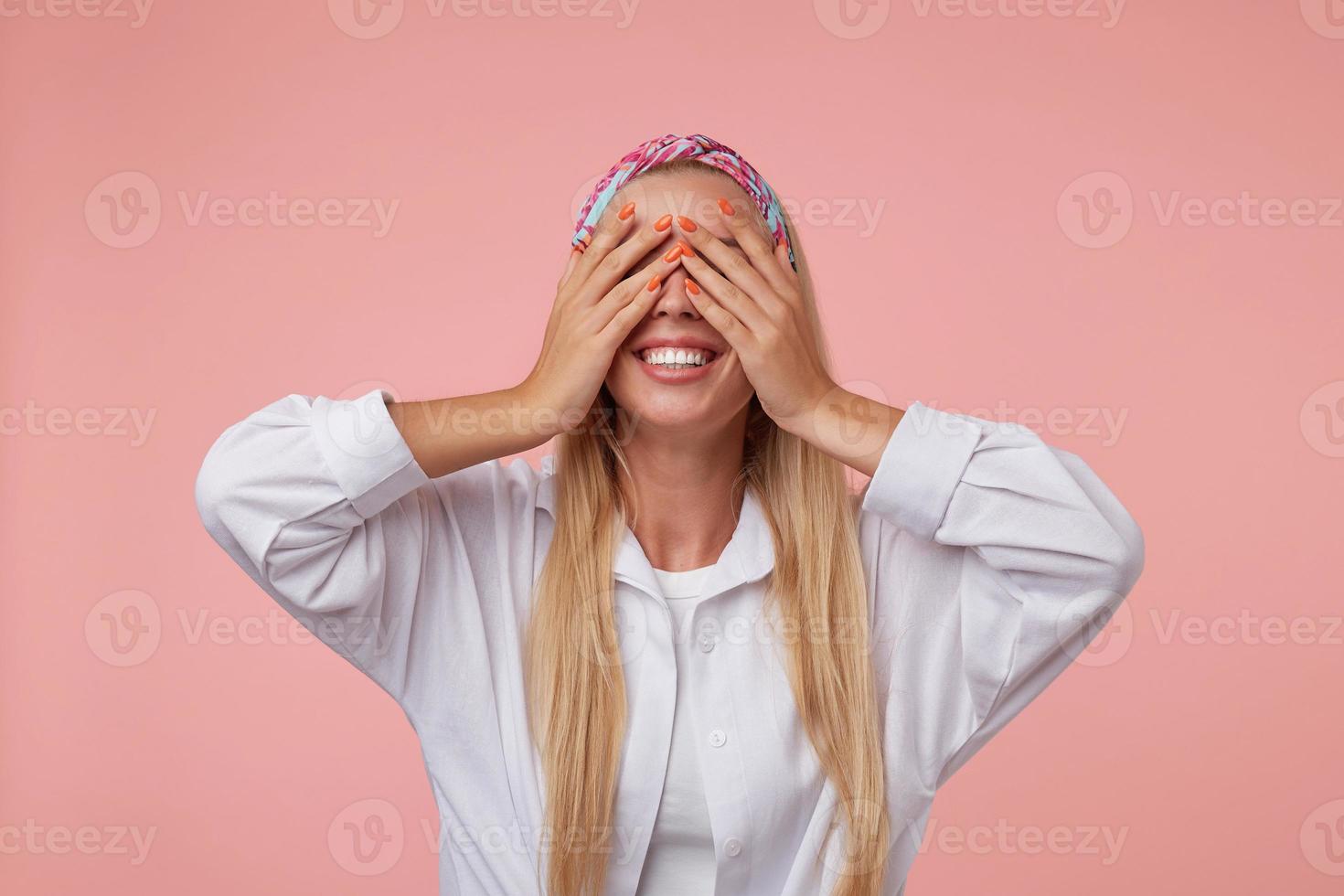 Happy young pretty blonde posing over pink background, standing with palms on her eyes and expecting pleasant surprise, wearing casual clothes photo