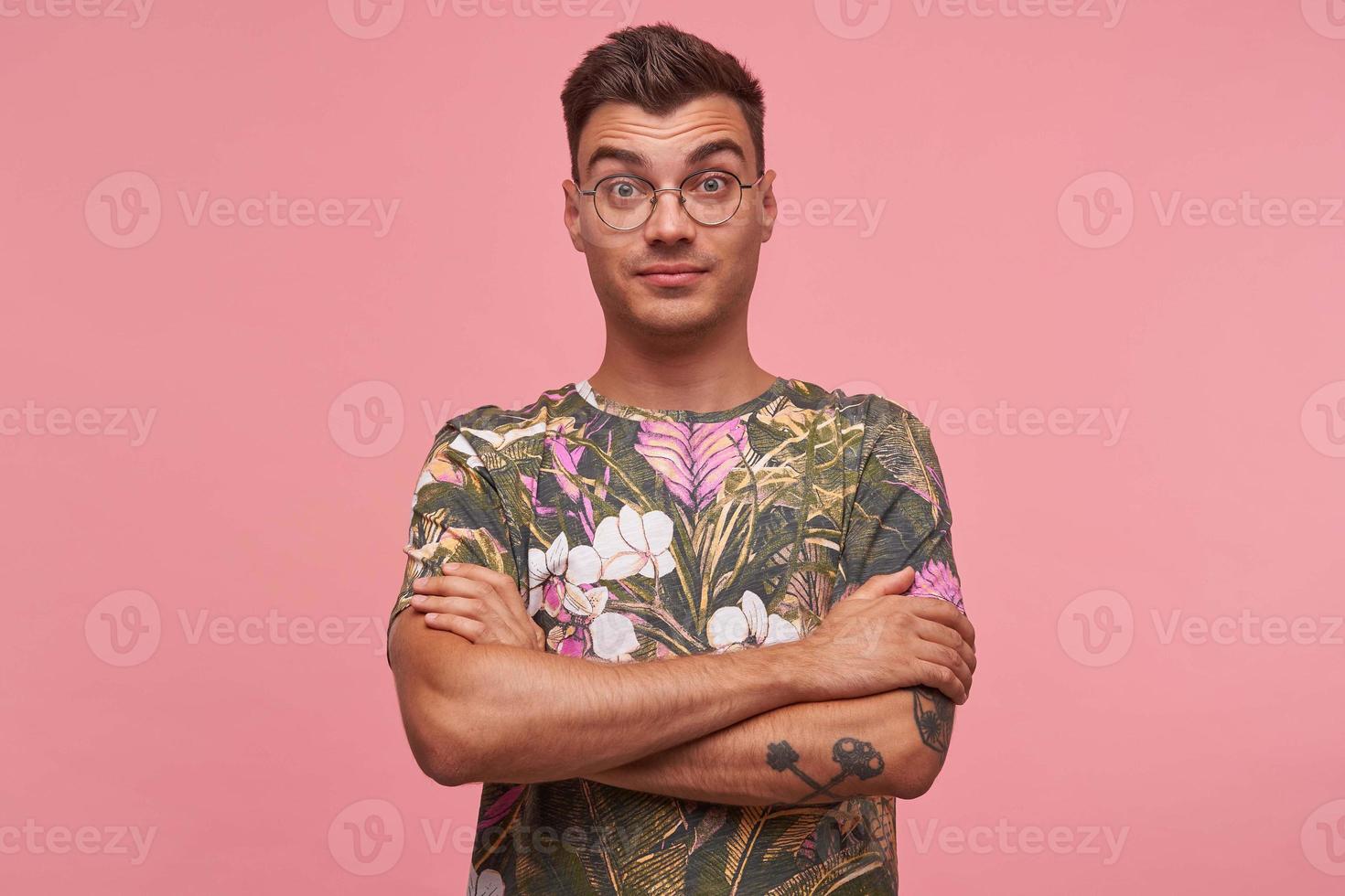 Studio portrait of surprised pretty young man with short haircut standing over pink background, looking puzzled to camera with crossed arms on his chest photo