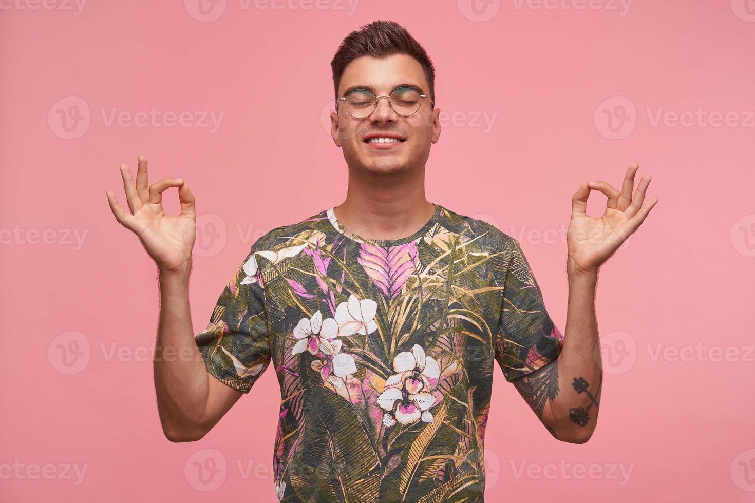 Happy smiling young man practicing indoor yoga exercise, meditating and breathing with eyes closed, showing mudra gesture, isolated on pink background photo