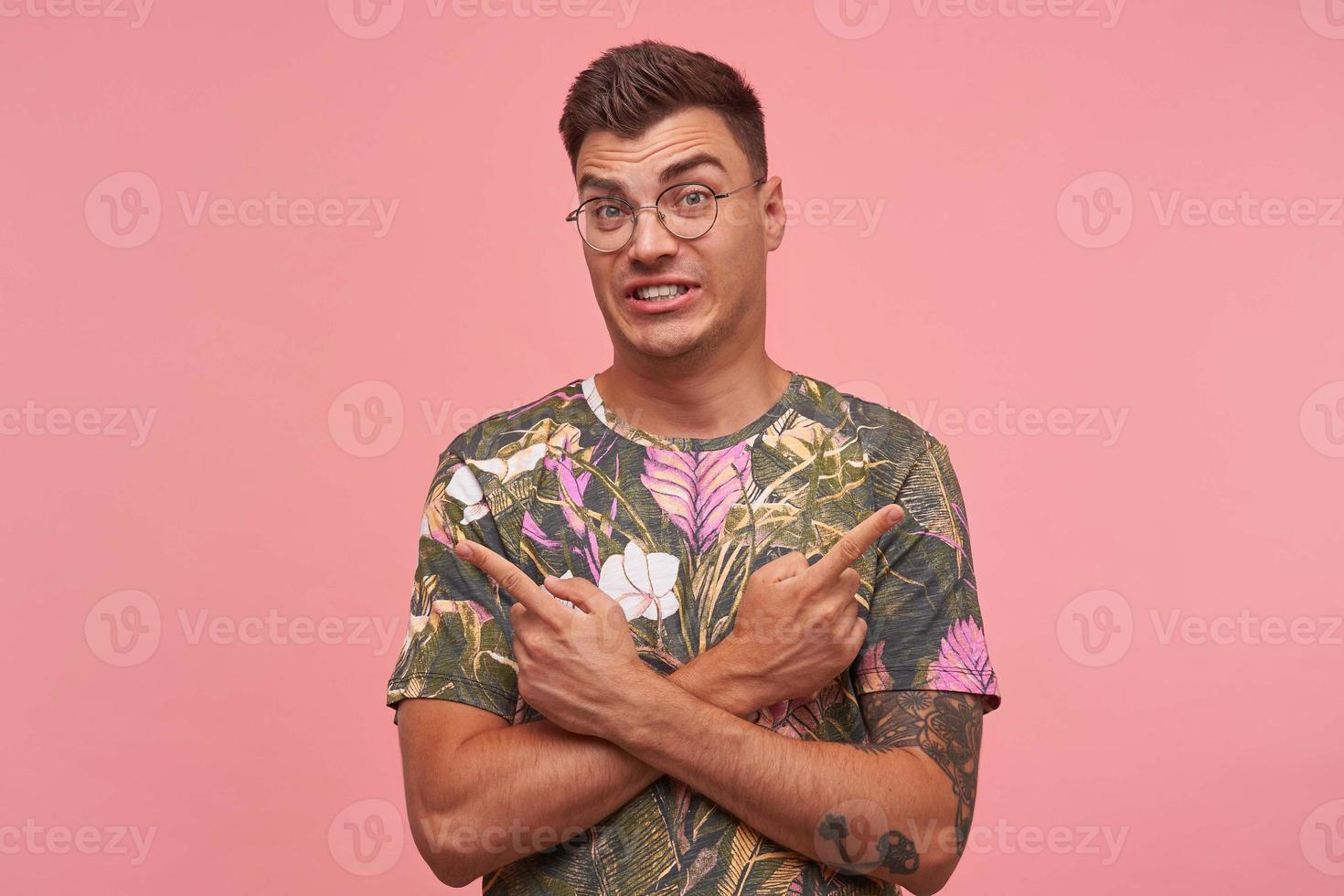 Confused young man in flowered t-shirt standing over pink background with crossed hands and pointing fingers to different sides, frowning and showing teeth photo
