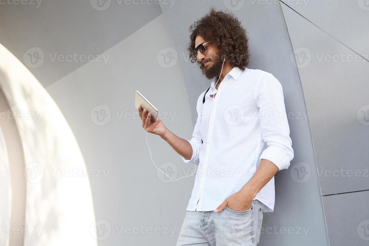 Young curly dark haired man with beard leaning on grey wall, posing over city background, having video calling and talking to somebody using headphones, smiling to tablet camera photo